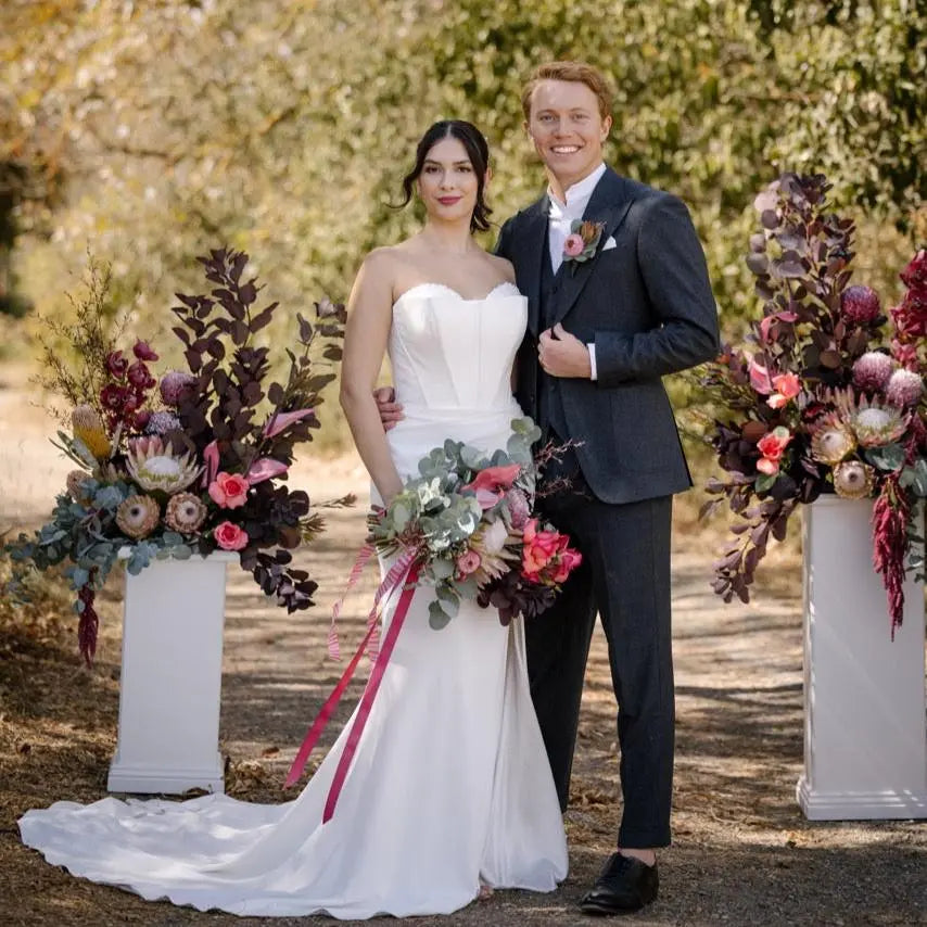 Photo of the Bride and Groom: Bride in the Allure wedding gown holding hands with the groom, showcasing the elegant fit-and-flare silhouette with a dramatic train in a garden setting.