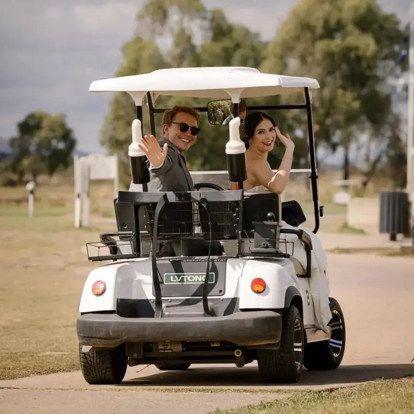 A bride and groom are seen from behind, smiling and waving as they ride in a golf cart. The bride is wearing a strapless white wedding gown, and the groom is dressed in a suit with sunglasses. The background features a natural outdoor setting with trees and a cloudy sky.