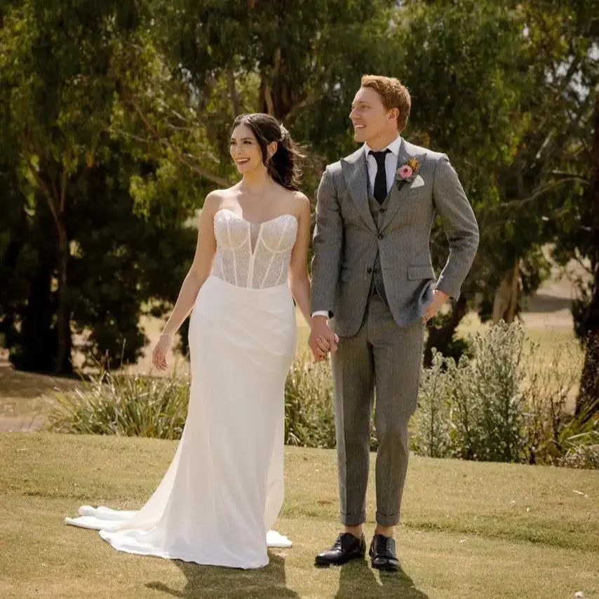 Bride in Kara Wedding Gown with beaded tulle overlay, corset bodice, and fit and flare skirt walking with groom outdoors