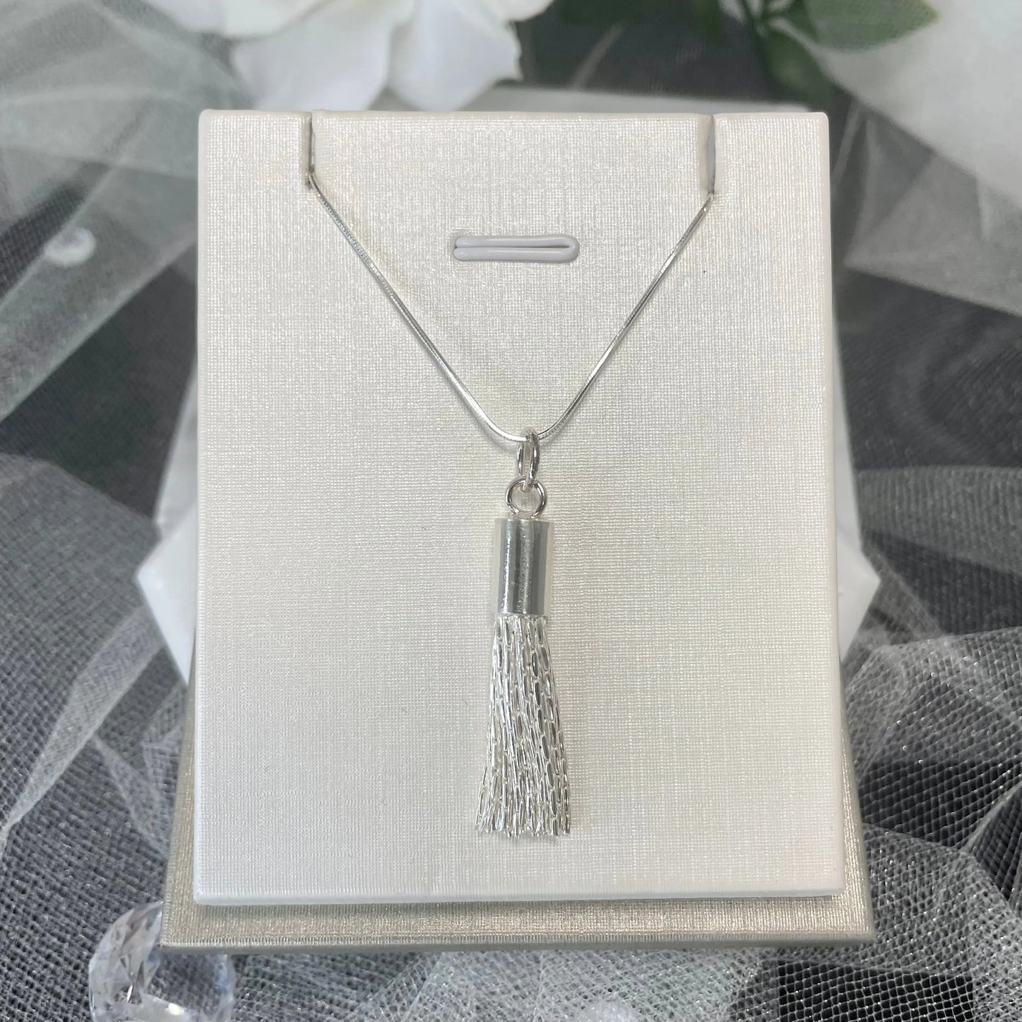 Elegant silver necklace with a tassel pendant on a minimalist display card, surrounded by soft white netting and decorative crystal elements, highlighting its sophisticated and modern design.