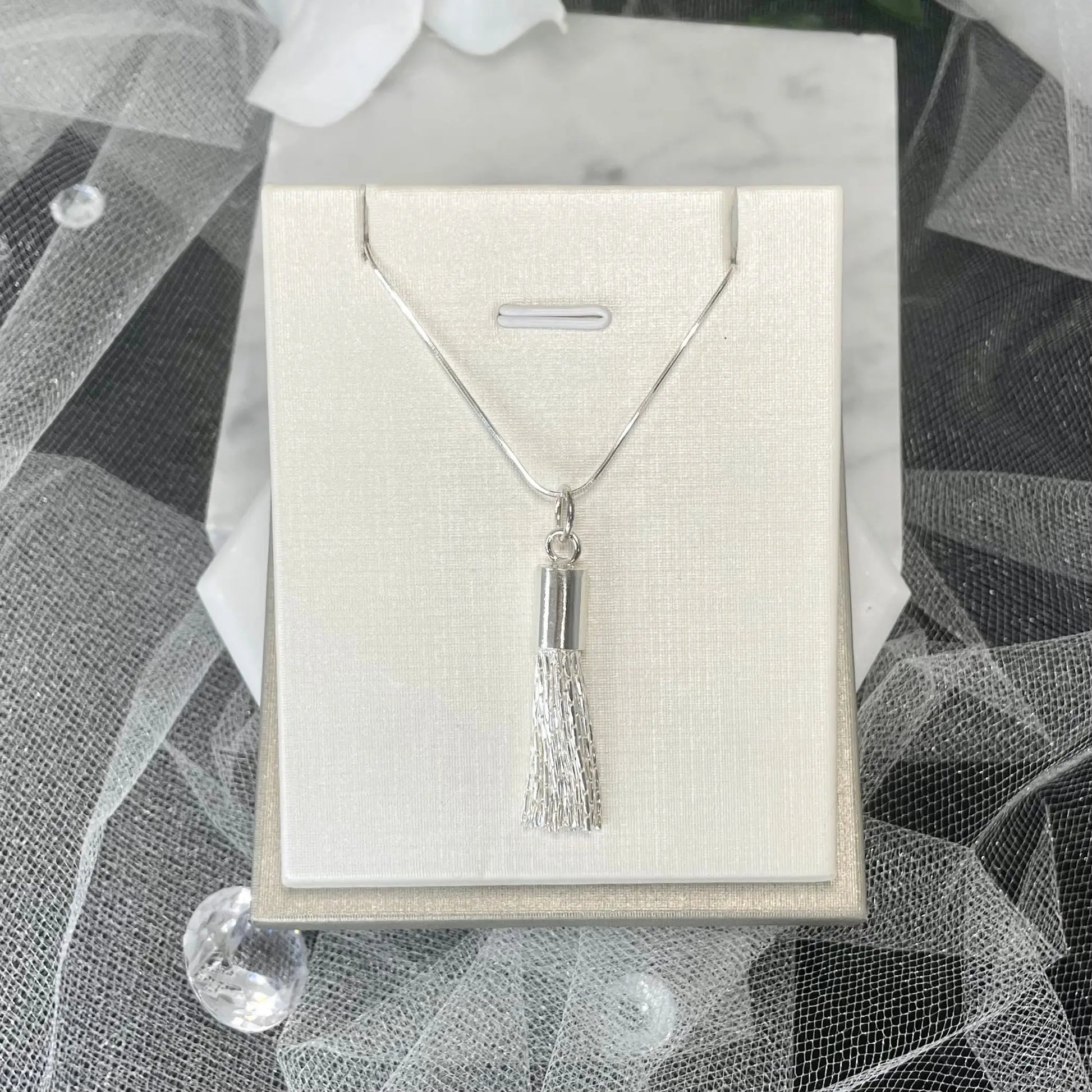 A silver necklace featuring a sleek, cylindrical pendant with fine, shimmering tassels displayed on a cream-colored jewelry card, set against a delicate backdrop of white tulle and sparkling crystal accents.
