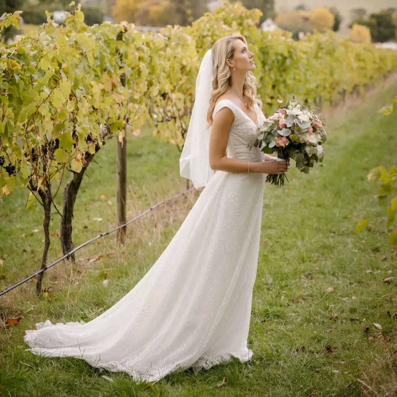 A radiant bride in the Tiana Wedding Gown smiles joyfully amidst a vineyard setting. The gown features an intricate full-lace overlay, scalloped neckline, and a romantic train, complemented by a delicate veil and a bouquet of soft roses and greenery