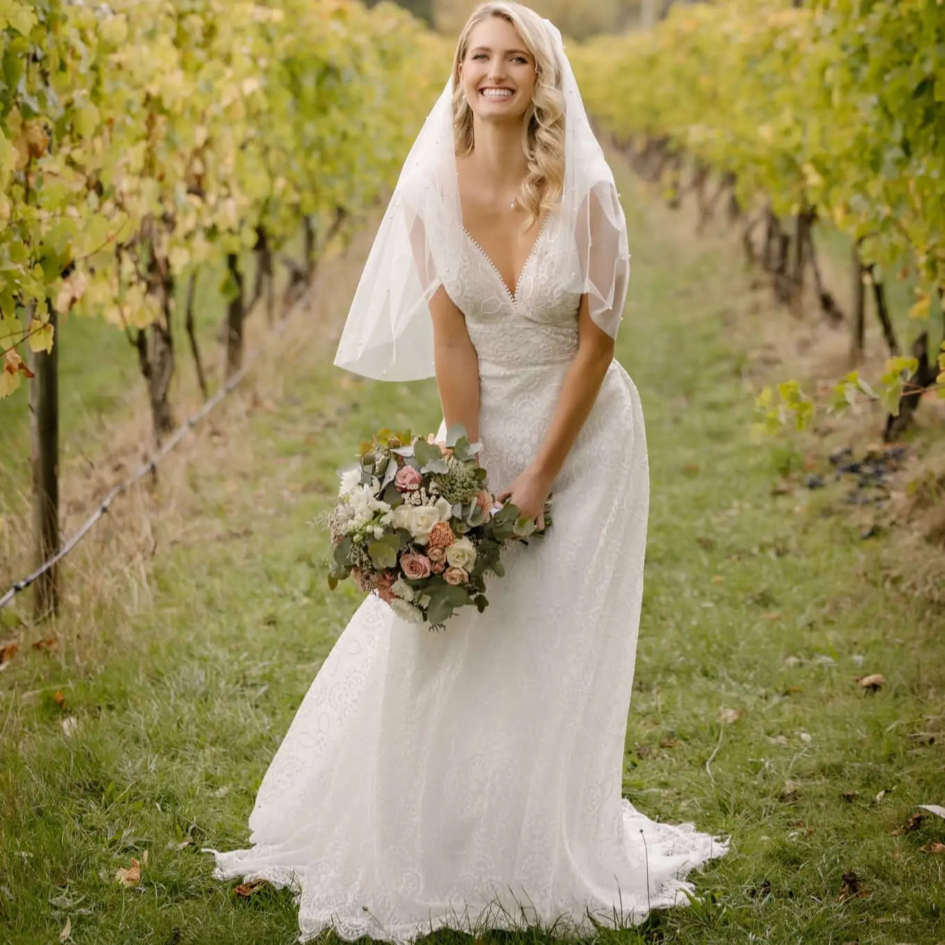 A radiant bride in the Tiana Wedding Gown smiles joyfully amidst a vineyard setting. The gown features an intricate full-lace overlay, scalloped neckline, and a romantic train, complemented by a delicate veil and a bouquet of soft roses and greenery