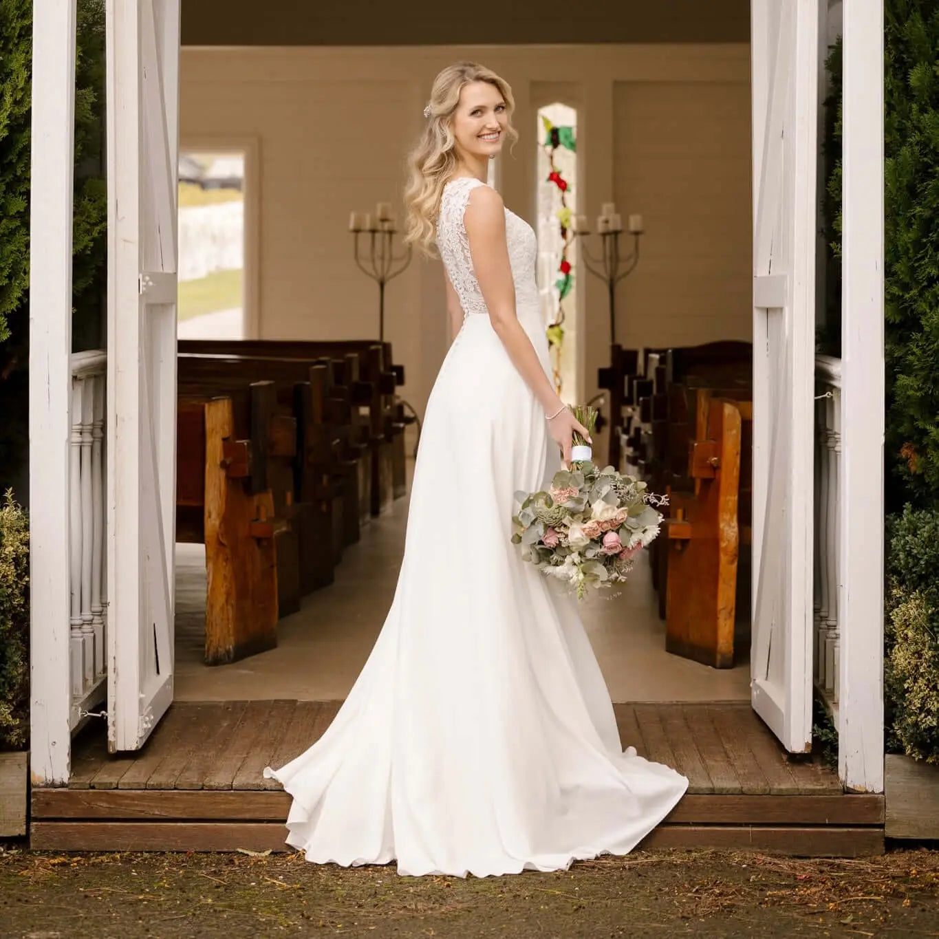 An elegant bride stands at the entrance of a charming chapel, poised in the Luna Wedding Gown. The dress features a plunging V-neckline with illusion mesh and form-fitting silhouette. The gown's smooth fabric cascades into a graceful chapel train, while the bride holds a bouquet of soft-colored flowers, completing her timeless bridal look.